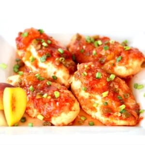 Four pieces of peach salsa chicken on plate with green onions, and fresh peach slices to the side.