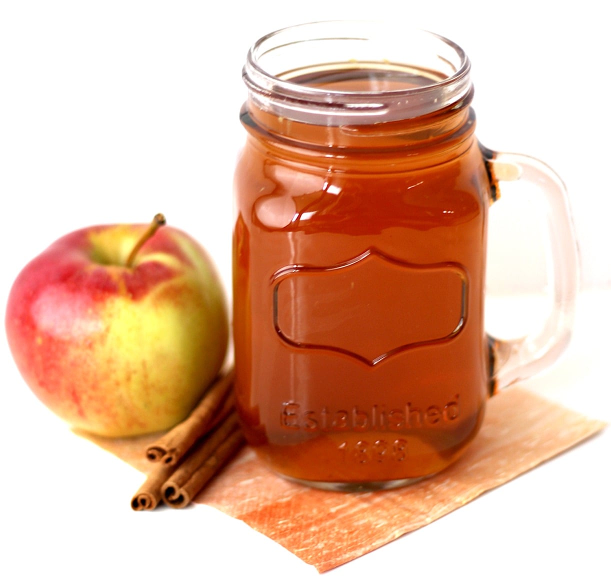 Spiced apple cider in mason jar mug with apple and cinnamon sticks to the side.