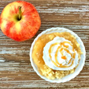 Apple caramel dump cake in small bowl with whip cream and caramel sauce on top, and apple to the side.