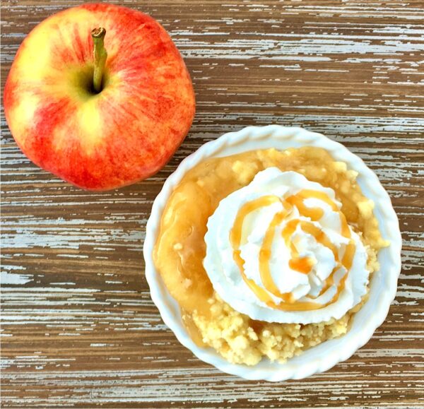 Apple caramel dump cake in small bowl with whip cream and caramel sauce on top, and apple to the side.