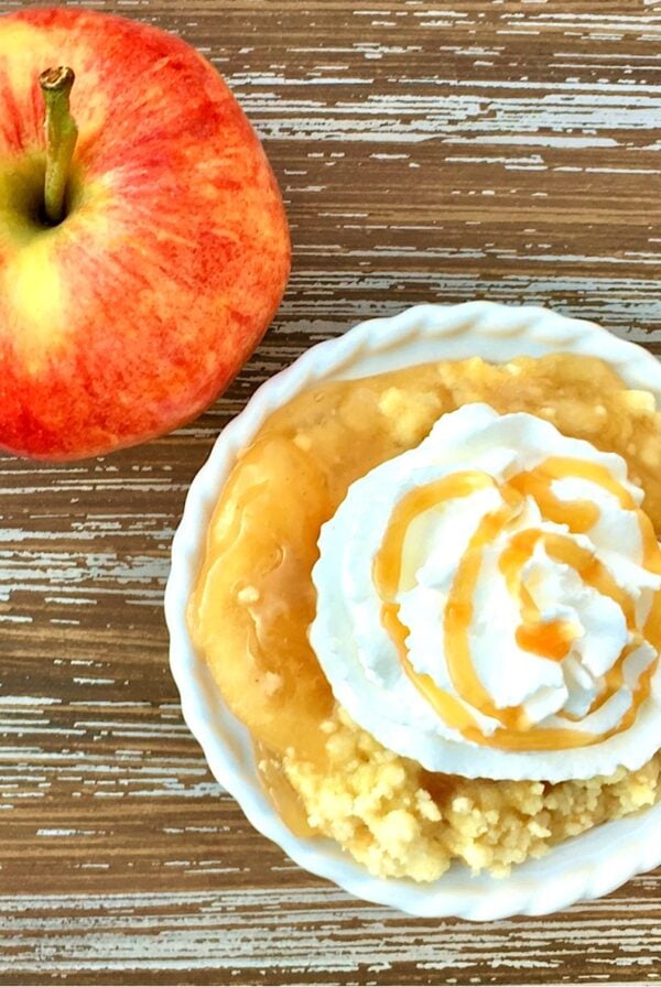 Apple caramel dump cake in small bowl with whip cream and caramel sauce on top, and apple to the side.