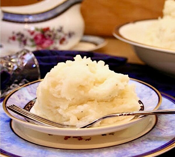 Fluffy mashed potatoes on plate with fork, and bowl of mashed potatoes in background.